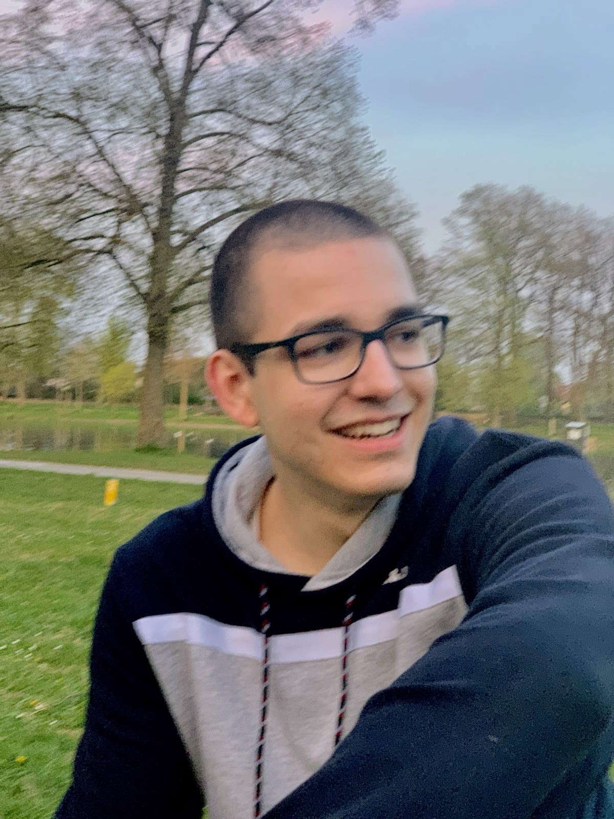 Portrait of Markus, a young man with short brown hair, black glasses and wearing a
                  hoodie, while sitting in a park 